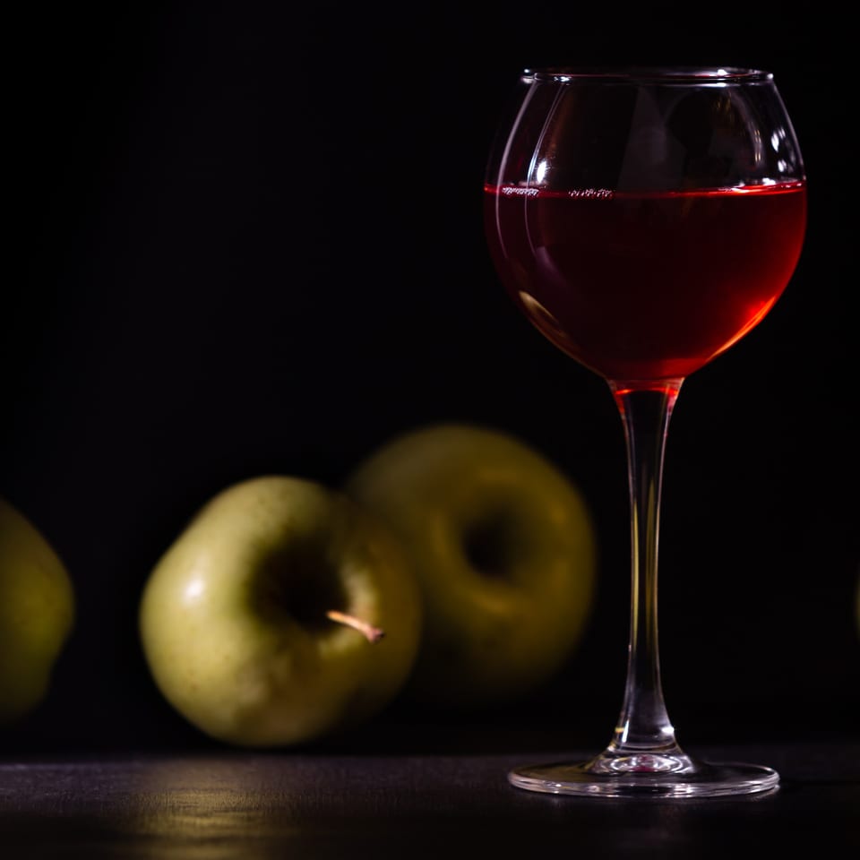 A glass of red wine stands beside several green apples on a dark surface.