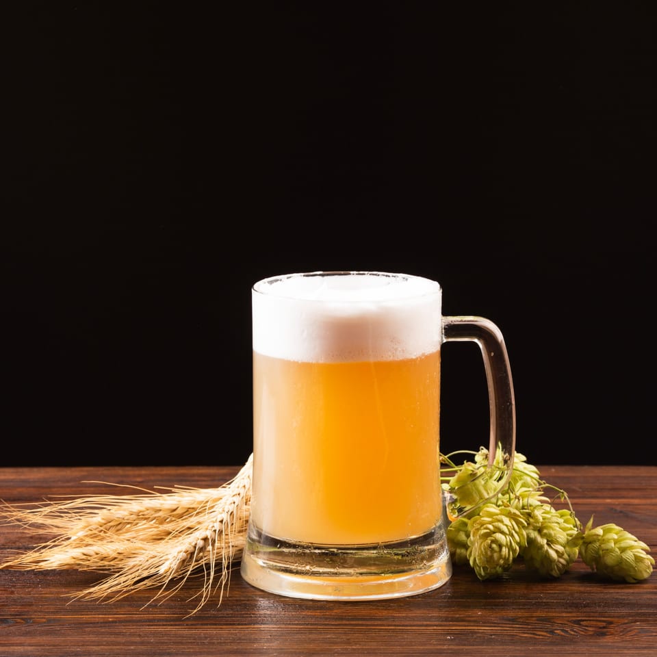 A frothy mug of beer sits on a wooden table with wheat stalks and hops.