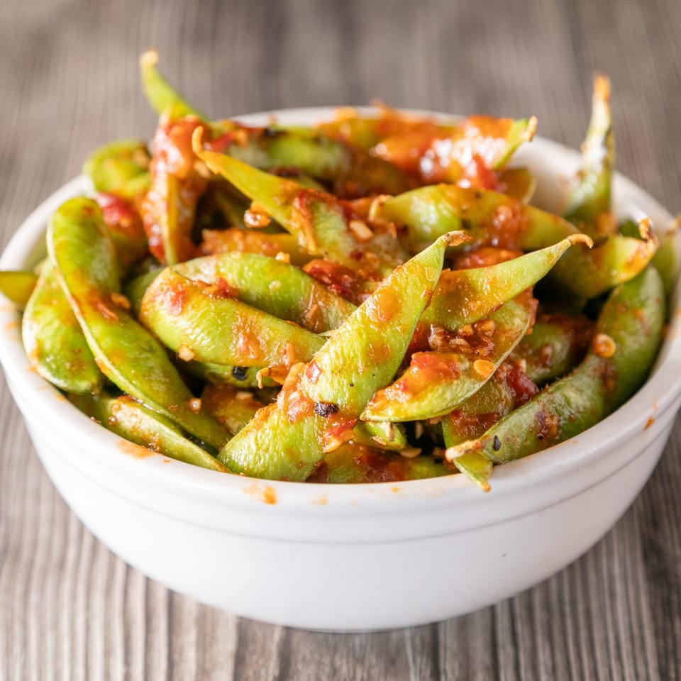 A white bowl filled with vibrant green edamame pods coated in a spicy red sauce.